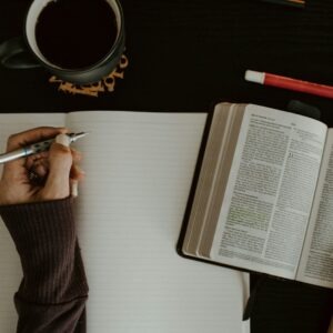 A hand holds a pen above a blank notebook, while the other hand gestures towards an open Bible. A cup of coffee sits nearby, creating a study atmosphere.