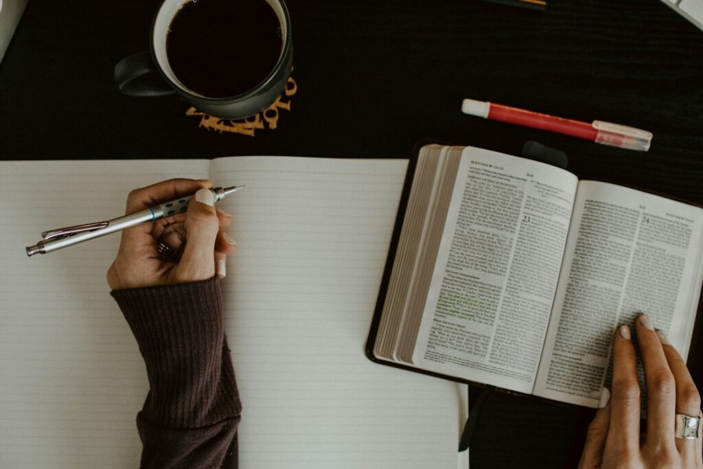 A hand holds a pen above a blank notebook, while the other hand gestures towards an open Bible. A cup of coffee sits nearby, creating a study atmosphere.