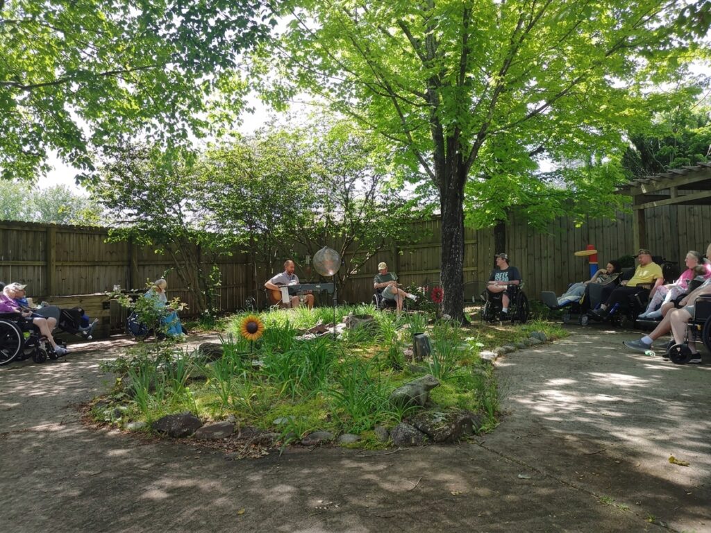 A music performer plays guitar in a serene garden, while residents in wheelchairs and on benches enjoy the outdoor entertainment.