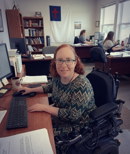 A woman in a wheelchair sits at a desk with a computer, smiling. Background features colleagues working in an office setting, showcasing a collaborative environment.